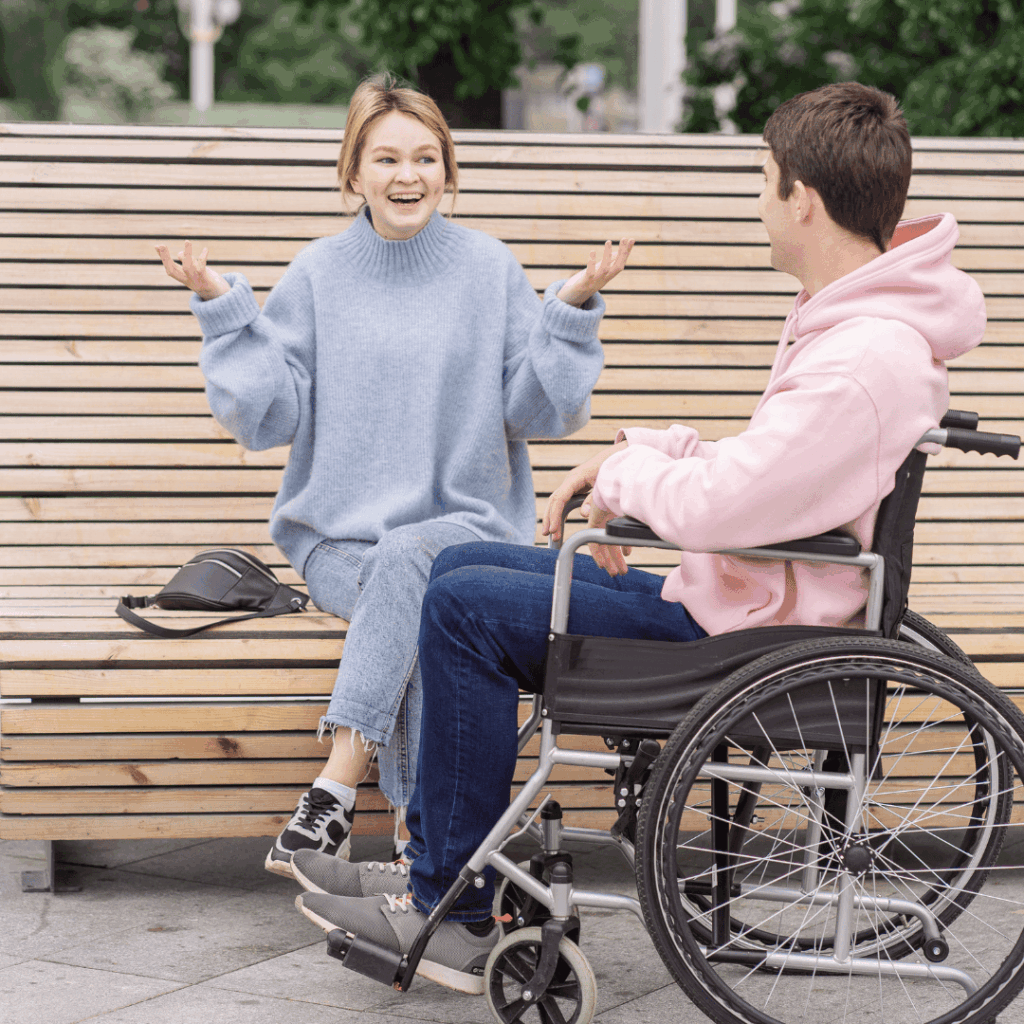 Kobieta siedząca na ławce rozmawia z mężczyzną na wózku inwalidzkim; oboje uśmiechnięci i zaangażowani w rozmowę.