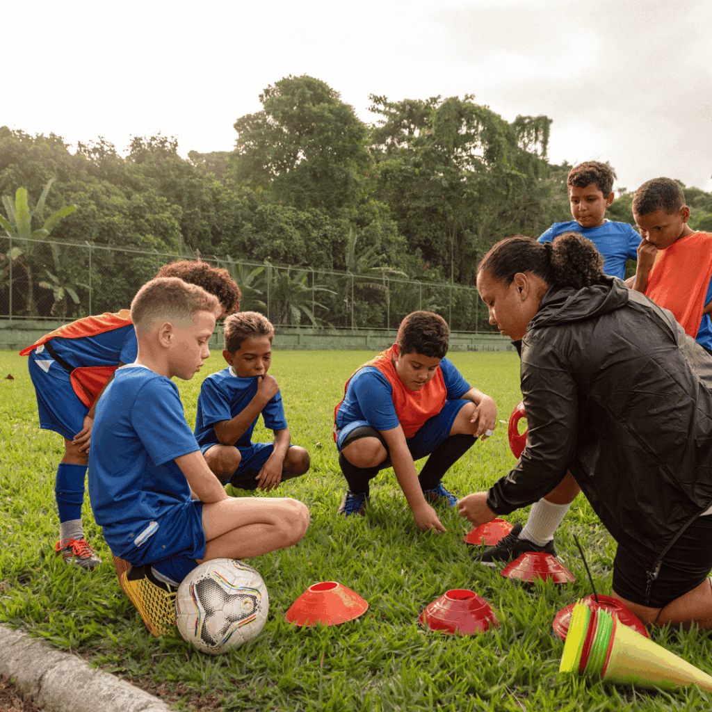 Trenerka tłumacząca ćwiczenie grupie dzieci w strojach piłkarskich siedzących na murawie boiska treningowego.